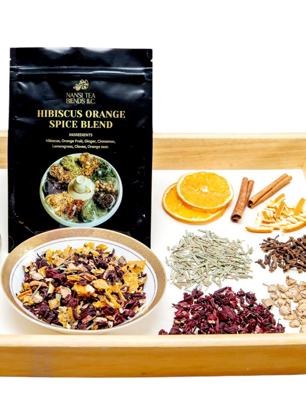 On a wooden tray is a pouch of Hibiscus Orange Spice Blend, a small glass bowl with loose leaf tea, surrounded by dry ingredients: hibiscus, orange slices, orange zest, cinnamon, ginger, cloves, and lemongrass.